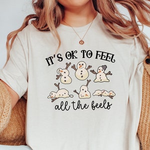 May include: Cream-colored t-shirt with the text "It's ok to feel all the feels" and a graphic of snowmen. The snowmen have stick arms and are wearing hats. The shirt is paired with blue jeans and a tan cardigan.