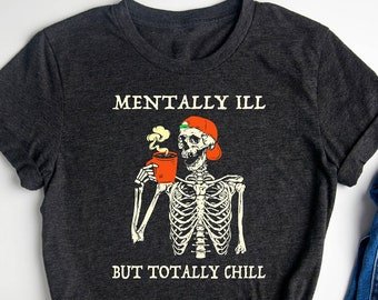 May include: A dark gray t-shirt with a skeleton wearing a Santa hat holding a cup of coffee. The text on the shirt reads "Mentally Ill But Totally Chill".