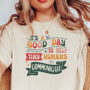 May include: A white t-shirt with a colorful graphic that says "It's a good day to help tiny humans communicate".