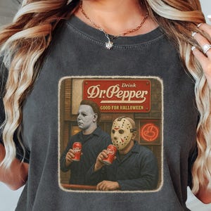 A woman wearing a t-shirt with a graphic of a man in a costume, holding a bottle of Dr Pepper, sitting on a bed.