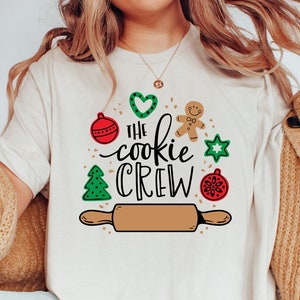 May include: White t-shirt with a Christmas cookie theme design. The design includes a rolling pin, a gingerbread man, a heart, a star, and ornaments. The text on the shirt reads "The Cookie Crew".