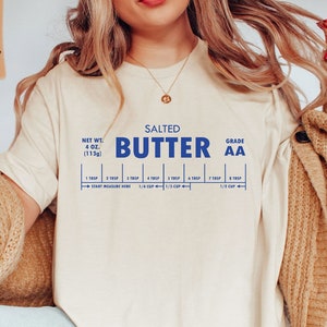 Puede incluir: Una camiseta blanca con un gr&aacute;fico azul de una barra de mantequilla con el texto "SALTED BUTTER GRADE AA" y una gu&iacute;a de medici&oacute;n para 1/4 taza, 1/3 taza y 1/2 taza.