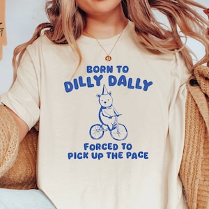 May include: A white t-shirt with blue text that reads "Born to Dilly Dally, Forced to Pick Up the Pace". A cartoon bear wearing a party hat is riding a bicycle.