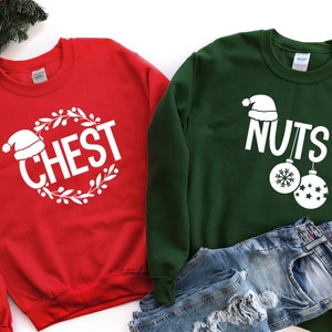 Peut inclure: Deux sweats à thème de Noël, rouge et vert, avec le texte "CHEST" et "NUTS" en blanc. Le sweat rouge a une couronne avec un chapeau de Père Noël sur le dessus. Le sweat vert a un chapeau de Père Noël et deux boules.
