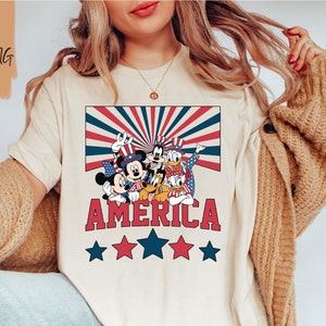 May include: A white t-shirt with a red, white, and blue graphic of Mickey Mouse, Donald Duck, Goofy, and Pluto wearing patriotic hats and holding American flags. The graphic is surrounded by a red, white, and blue striped background with the word "AMERICA" in red letters and five stars below.