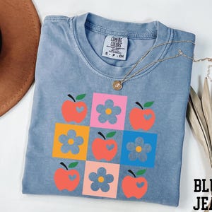 a blue t-shirt with a colorful graphic design of apples and flowers, along with a necklace and a hat.