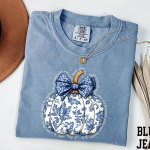 A blue t-shirt with a white pumpkin design and a blue bow, along with a brown hat and some leaves, are displayed on a white surface.