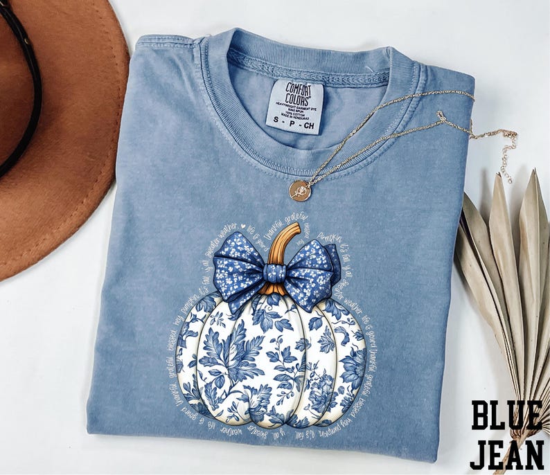 A blue t-shirt with a white pumpkin design and a blue bow, along with a brown hat and some leaves, are displayed on a white surface.