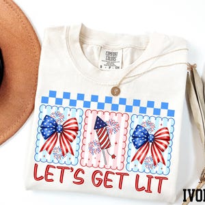 a white t-shirt with a patriotic design, including a fireworks display and a ribbon with the text "Let's Get Lit". The t-shirt is placed on a white background, along with a brown hat and a fan.