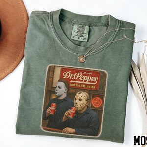 A green t-shirt with a graphic design featuring two men wearing masks, one holding a can of Dr. Pepper, and the other holding a can of Diet Coke.