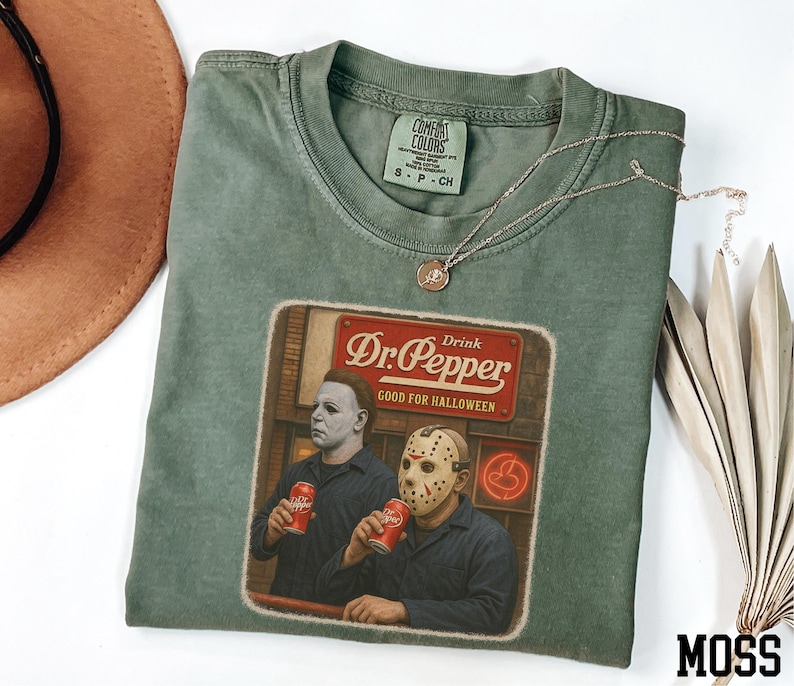 A green t-shirt with a graphic design featuring two men wearing masks, one holding a can of Dr. Pepper, and the other holding a can of Diet Coke.