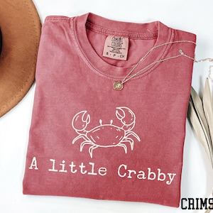 A red t-shirt with a white crab design and the text "A little Crabby" printed on it, along with a necklace and a hat.
