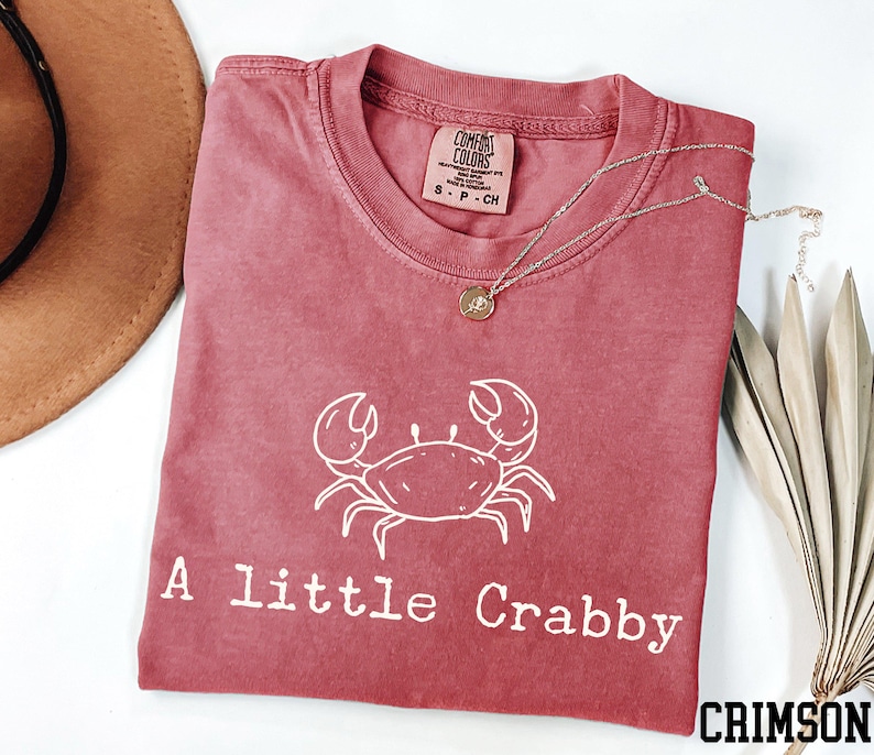 A red t-shirt with a white crab design and the text "A little Crabby" printed on it, along with a necklace and a hat.