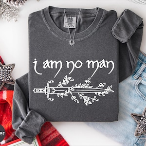 May include: A dark gray long-sleeve shirt with the text "i am no man" in white script. Below the text is a white illustration of a sword with leaves. The shirt is folded on a white surface with a red plaid scarf and jeans.