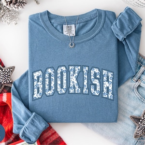 May include: A blue jean long-sleeve shirt with the word "BOOKISH" in white block letters filled with a blue floral pattern. A silver necklace with a sun pendant is on the shirt. The shirt is folded on a white surface.