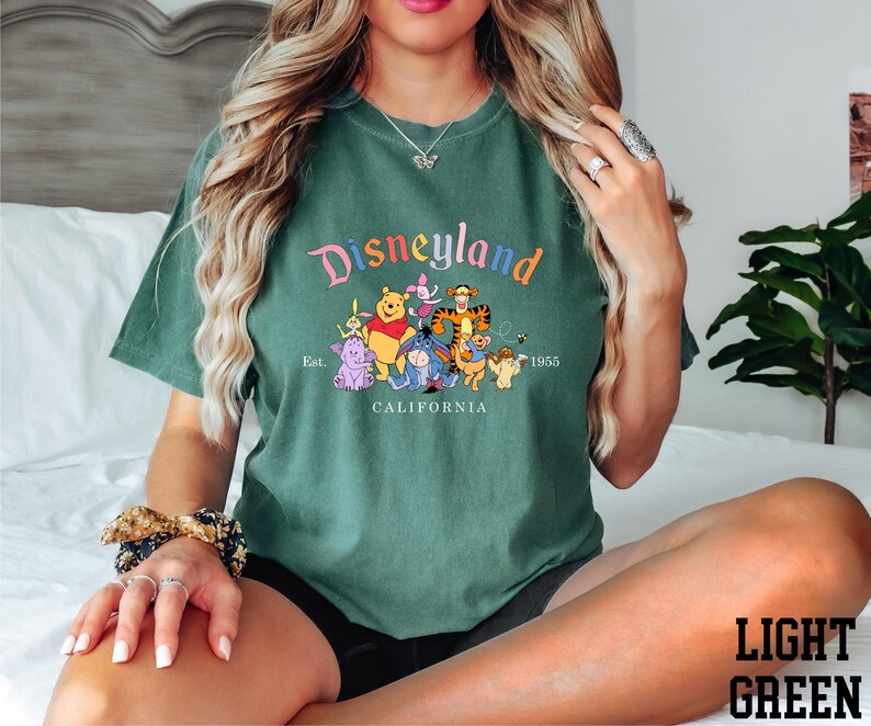 A woman wearing a green t-shirt with a Disneyland graphic and text, sitting on a bed with a potted plant in the background.
