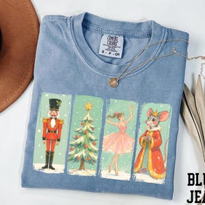 May include: A light blue Comfort Colors t-shirt featuring a Nutcracker-themed graphic. The graphic includes a nutcracker, Christmas tree, ballerina, and mouse king. A gold necklace and a brown hat are also visible.