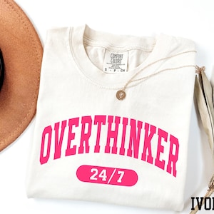a white t-shirt with the text "OVERTHINKER" in pink letters, along with the number "24/7" below it. The t-shirt is laid out on a white surface, with a brown hat and a fan visible in the background.