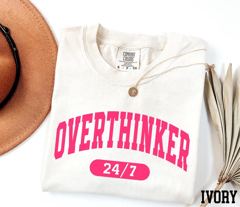 a white t-shirt with the text "OVERTHINKER" in pink letters, along with the number "24/7" below it. The t-shirt is laid out on a white surface, with a brown hat and a fan visible in the background.
