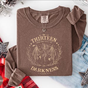 May include: Espresso-colored long-sleeve shirt with the text "the THIRTEEN from now until the DARKNESS claims us" in a circular design. The shirt has a crew neck and a silver necklace. A pair of blue jeans and star-shaped ornaments are also visible.