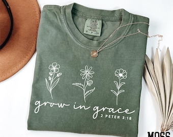 May include: Moss green t-shirt with white floral illustrations and the phrase "grow in grace" printed on the front. A gold necklace rests on the shirt. The shirt is a Comfort Colors brand.