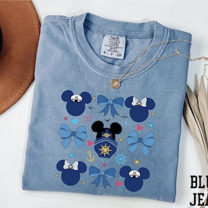 May include: A light blue t-shirt with a crew neck featuring a graphic design of Mickey Mouse heads, bows, and nautical elements. The shirt has the text "BLUE JEAN" in the bottom right corner. A gold necklace is draped across the shirt.