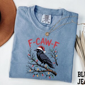 A blue t-shirt with a graphic design of a black crow wearing a Santa hat, surrounded by Christmas lights and branches.