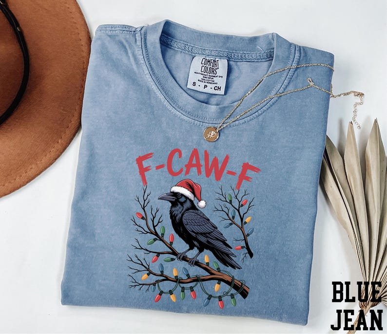 A blue t-shirt with a graphic design of a black crow wearing a Santa hat, surrounded by Christmas lights and branches.