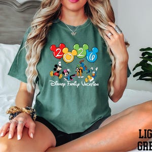 A woman wearing a green t-shirt with a Disney character graphic and the text "Disney Family Vacation" is sitting on a bed.
