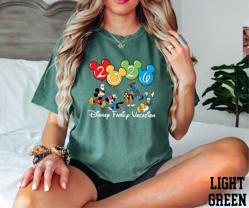 A woman wearing a green t-shirt with a Disney character graphic and the text "Disney Family Vacation" is sitting on a bed.
