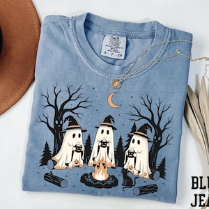 Peut inclure: Un t-shirt bleu avec un motif représentant trois fantômes portant des chapeaux de sorcière assis autour d'un feu de camp dans une forêt. Les fantômes tiennent des tasses et un chat noir est assis à proximité. Étiquette Comfort Colors S P CH.