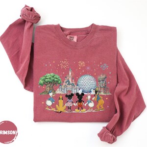 A pink long-sleeved shirt with a Disney-themed graphic design featuring Mickey Mouse, Donald Duck, Goofy, and other iconic Disney characters, along with a castle and fireworks in the background.
