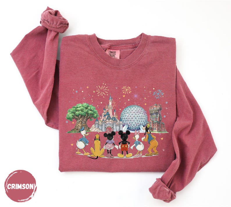 A pink long-sleeved shirt with a Disney-themed graphic design featuring Mickey Mouse, Donald Duck, Goofy, and other iconic Disney characters, along with a castle and fireworks in the background.