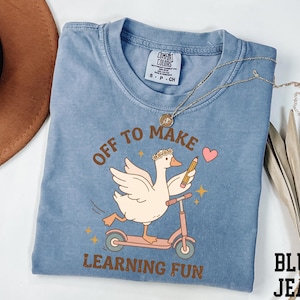 May include: Blue jean-colored t-shirt with a graphic of a goose riding a pink scooter, holding a pencil and a heart. The text on the shirt reads "Off to Make Learning Fun". A gold necklace is draped across the shirt.