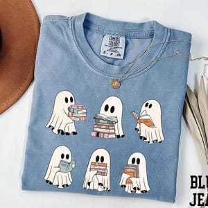 May include: A light blue Comfort Colors t-shirt featuring a design of cartoon ghosts holding and reading books. The ghosts are white with black eyes and feet. A gold necklace with a pendant is on the shirt. The words "BLUE JEAN" are in the bottom right corner.