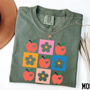 a green t-shirt with a colorful graphic design of apples and flowers, along with a brown hat and some dried leaves.
