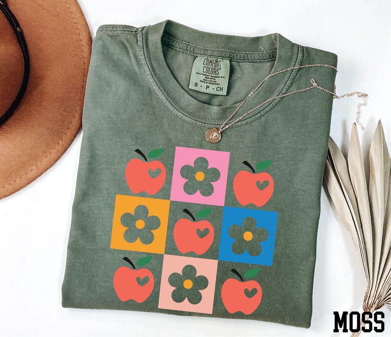 a green t-shirt with a colorful graphic design of apples and flowers, along with a brown hat and some dried leaves.