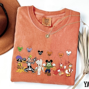 a red t-shirt with a cartoon character design, including Mickey Mouse, Goofy, Donald Duck, Minnie Mouse, and other Disney characters, along with balloons. The t-shirt is placed on a white surface, and there is a brown hat and a fan nearby.