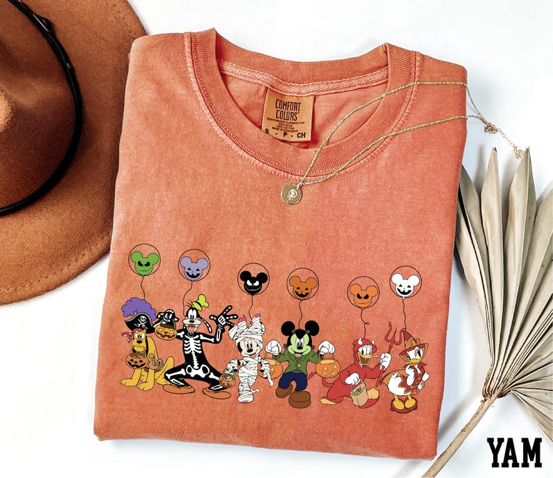 a red t-shirt with a cartoon character design, including Mickey Mouse, Goofy, Donald Duck, Minnie Mouse, and other Disney characters, along with balloons. The t-shirt is placed on a white surface, and there is a brown hat and a fan nearby.