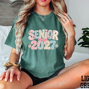A woman wearing a green t-shirt with the text "SENIOR 2027" printed on it, sitting on a bed.