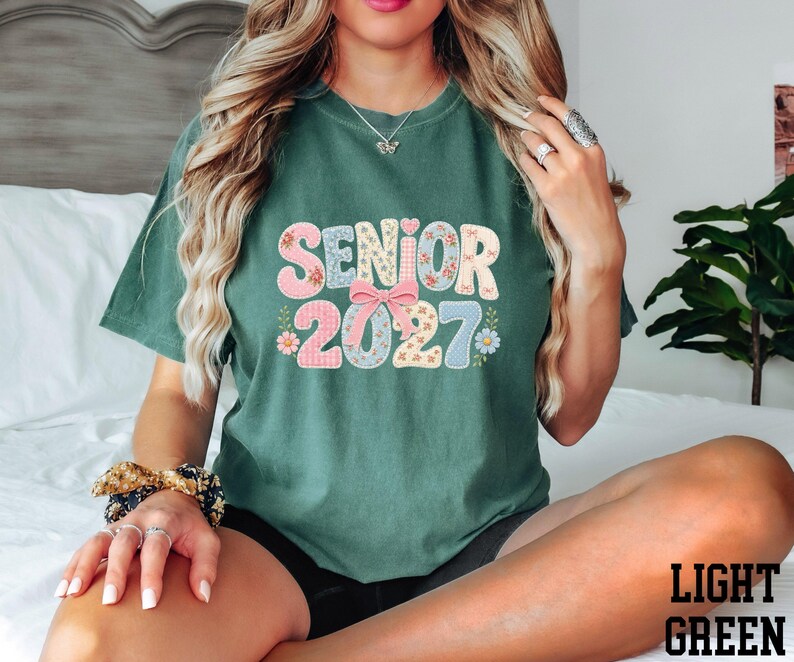 A woman wearing a green t-shirt with the text "SENIOR 2027" printed on it, sitting on a bed.