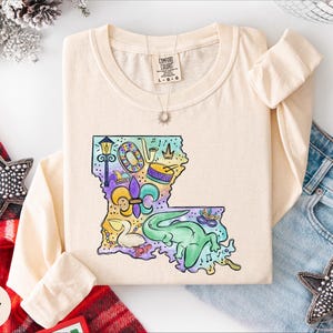 a beige long-sleeved t-shirt with a colorful graphic design depicting a map of Louisiana, along with various decorative elements such as stars and a crown. The t-shirt is laid out on a white surface, surrounded by other clothing items like jeans and a red scarf.