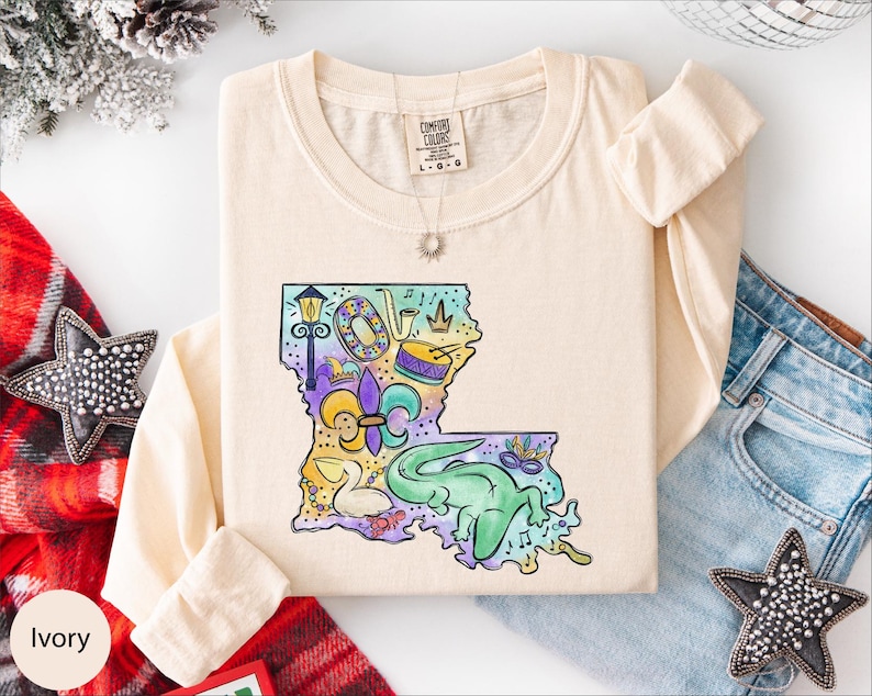 a beige long-sleeved t-shirt with a colorful graphic design depicting a map of Louisiana, along with various decorative elements such as stars and a crown. The t-shirt is laid out on a white surface, surrounded by other clothing items like jeans and a red scarf.