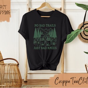 Może przedstawiać: Czarny T-shirt z zielonym nadrukiem przedstawiającym dw&oacute;ch turyst&oacute;w w lesie z tekstem "No Bad Trails Just Bad Knees".