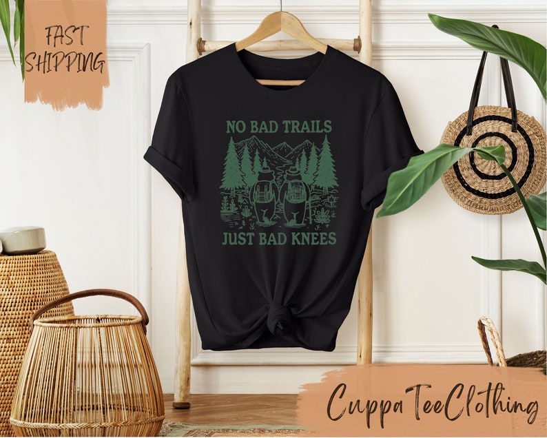 Może przedstawiać: Czarny T-shirt z zielonym nadrukiem przedstawiającym dw&oacute;ch turyst&oacute;w w lesie z tekstem "No Bad Trails Just Bad Knees".