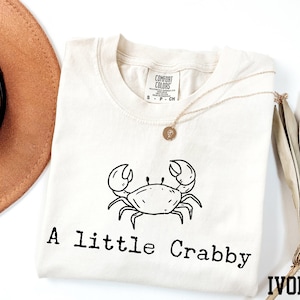 A white t-shirt with a black crab design and the text "A Little Crabby" printed on it, along with a necklace and a hat.