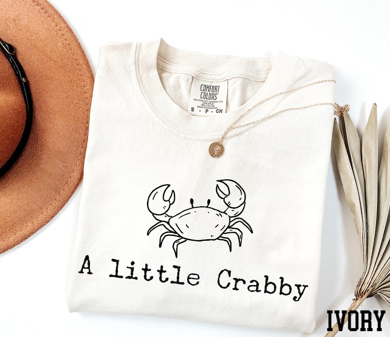 A white t-shirt with a black crab design and the text "A Little Crabby" printed on it, along with a necklace and a hat.