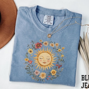 A blue t-shirt with a sun design and a floral wreath, along with a brown hat and a necklace, are arranged on a white surface.