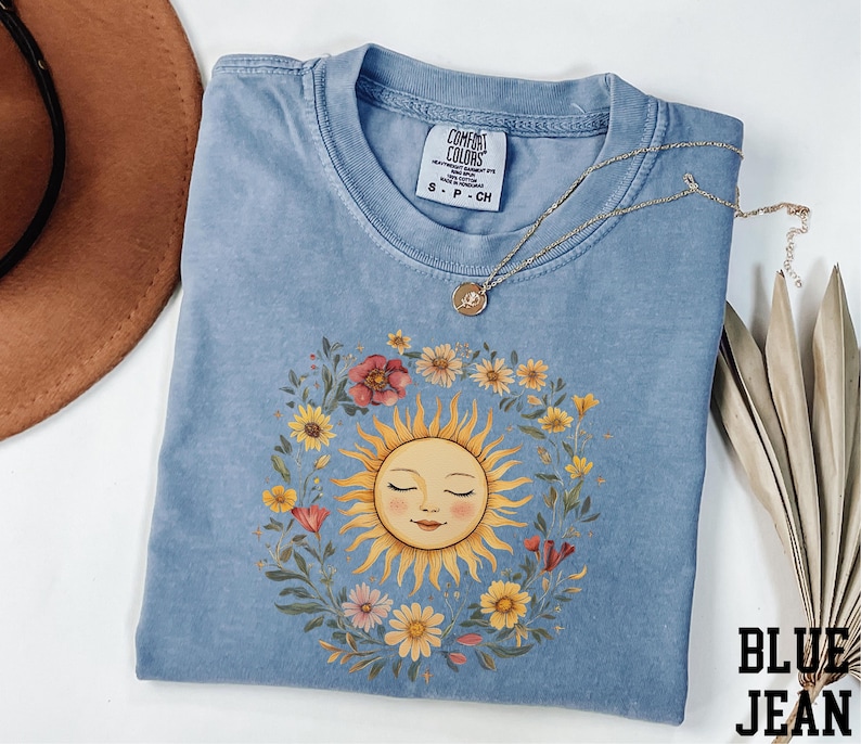A blue t-shirt with a sun design and a floral wreath, along with a brown hat and a necklace, are arranged on a white surface.
