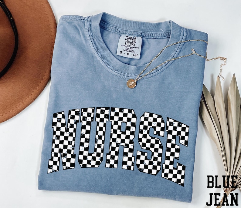 a blue t-shirt with a checkered pattern and the word "NURSE" printed on it. The t-shirt is laid out on a white surface, along with a brown hat and some leaves.